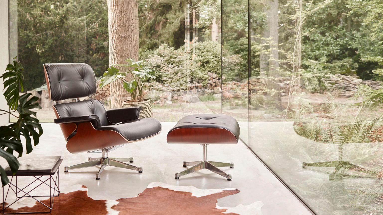 Vitra Eames Lounge Chair mit Ottoman aus schwarzem Leder und Holzfurnier, platziert vor bodentiefen Fenstern mit Blick ins Grüne – exklusiver Designklassiker bei Loeser Braunschweig