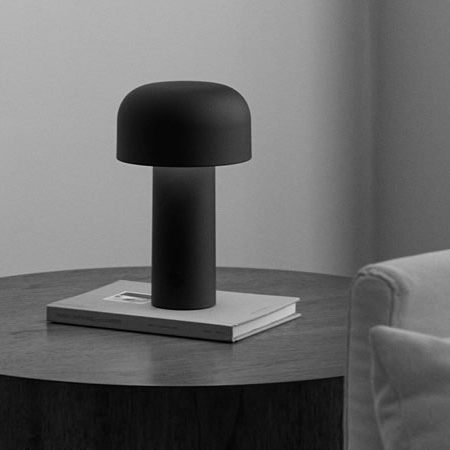 Flos Bellhop Tischleuchte in mattem Schwarz auf rundem Holztisch neben Sofa – puristisches Lichtdesign mit weicher Formensprache und moderner Eleganz