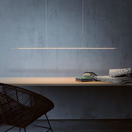 Elegante Gera Pendelleuchte über schlichtem Holztisch vor grauer Betonwand – minimalistisches Lichtdesign mit feiner Linienführung und atmosphärischer Ausleuchtung