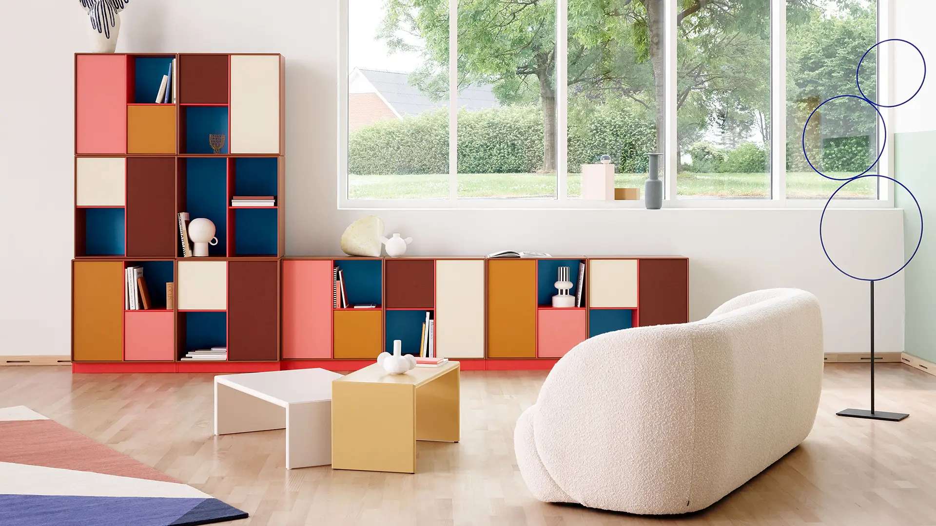 Modernes Wohnzimmer mit farbenfrohen Montana Regalsystemen in Rot-, Gelb- und Blautönen, gemütlichem Sofa und minimalistischer Dekoration vor großer Fensterfront
